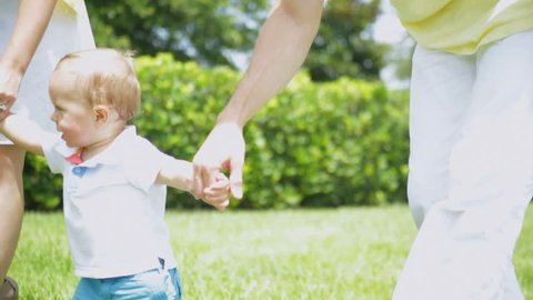 Happy Little Caucasian Boy Practicing Walking Park Grass - Smiling male Caucasian toddler child holding young parents hands slowly walking barefoot by on grass outside park