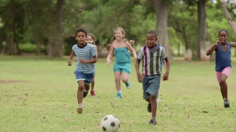 Young people, happy friends and ball sport, kids and children, multiethnic group of  boys and girls playing european football, american soccer in city park. Summer camp fun. Slow motion.