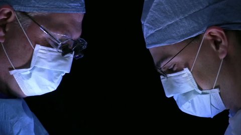 Two surgeons working together in front of a black background.  Lit from below.