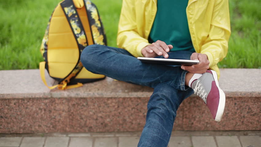 Young guy with original appearance sitting in the park and enjoys tablet
