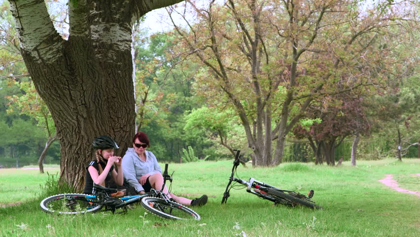Mother and teen daughter sitting under tree in the park with bicycles