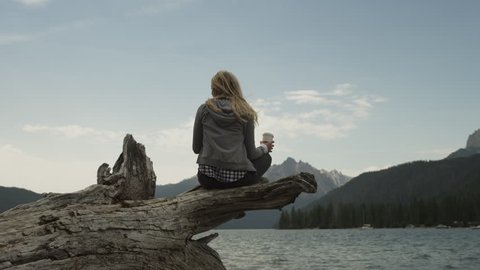 Medium shot of young woman drinking coffee on log at lake / Redfish Lake, Idaho, United States