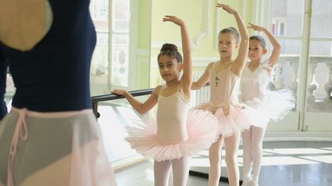 Female Ballet Dancer stands before a trio of young Ballerinas demonstrating the movements and encouraging leg and arm extension
