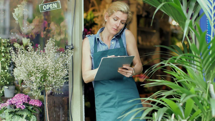 A Florist speaks on the phone, jotting notes eagerly as she leans against the doorway of her shop Royalty-Free Stock Footage #6441122