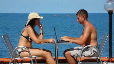 man using notebook and woman in swimsuit with cell phone sits at table 