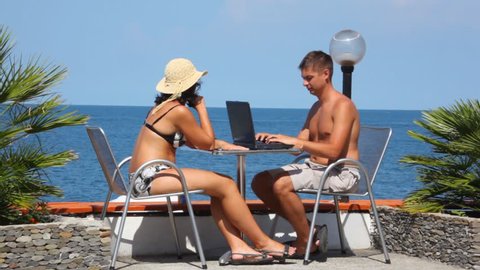 man using notebook and woman with cell phone sits at table, sea in background 