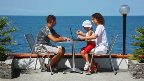 family of three persons with notebook sits at table, sea in background 