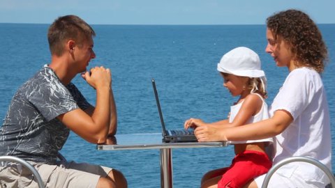 happy family of three persons with notebook sits at table, sea in background 
