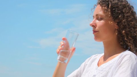 beautiful young woman drinks water from glass, blue sky in background 