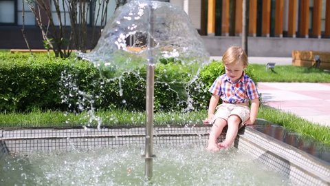 Little handsome boy sits in summer park down feet in fountain