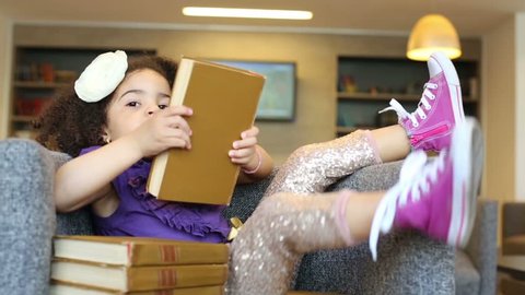 Little girl sits in armchair with stack of books and reads