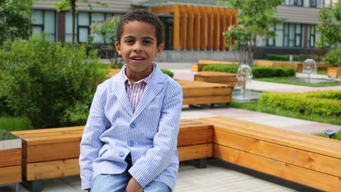 Happy cute boy in jacket sit on bench in summer park near business center