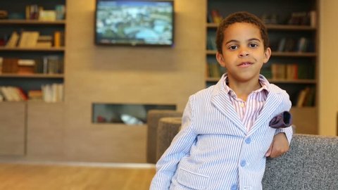 Cute boy in jacket stands near armchair and poses in business center