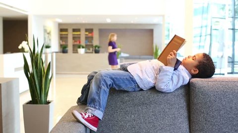 Curly boy in jacket and jeans lies on sofa and reads in business center