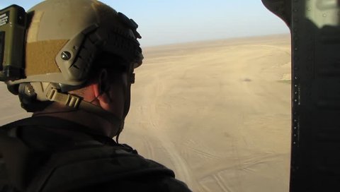 US soldier in helmet and combat armor sits in open doorway of military helicopter flying over desert in middle east