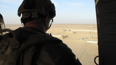 US soldier in helmet and combat armor sits in open doorway of military helicopter flying over desert base in middle east