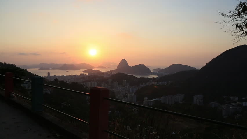 A man with a soccer ball walks into the frame of a wide shot of Sugarloaf mountain and the surrounding buildings in the city of Rio de Janeiro at sunset