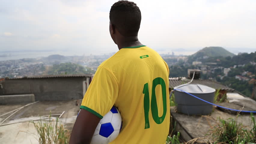 A kid in a soccer jersey stands with a ball under his arm while looking out at the city of Rio de Janeiro