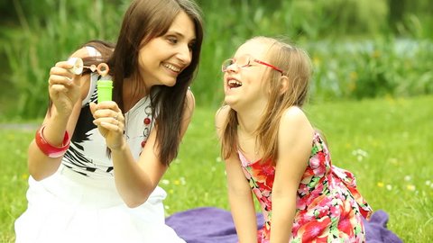Summer. Mother with daughter having fun. Young woman and girl child kid blowing soap bubbles on the meadow or in the park.