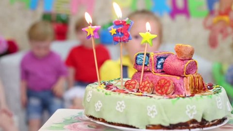 Birthday cake with three candles is on table and five kids expect treats at children party. Focus on cake