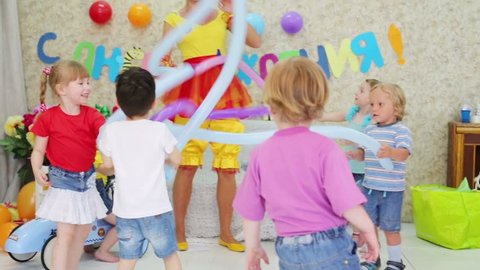 Five happy kids and facilitator play with long balloons at funny children party. Inscription Happy Birthday on wall