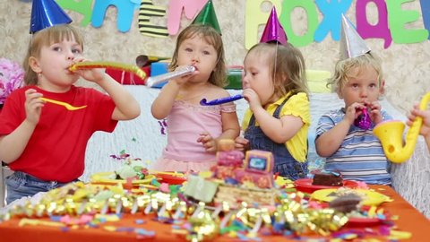Five little kids sit at red table with cake and blow in multicolor party blowers at birthday party. Inscription Happy Birthday on wall