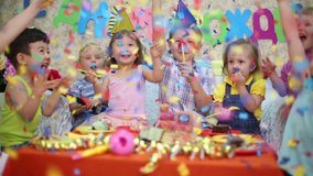 Seven little kids sit at red table with cake and throw confetti at birthday party. Inscription Happy Birthday on wall - Powered by Shutterstock - Get 15% off with code: PIKWIZARD15
