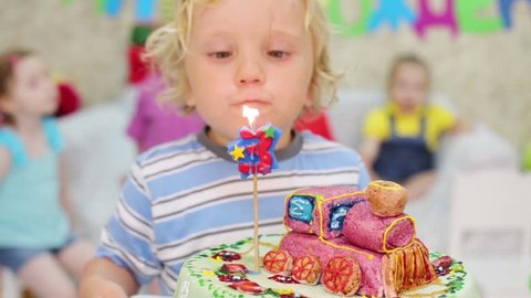 Little cute boy blows out one candle on cake and other kids look at he at children party. Focus on boy