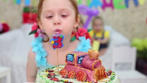 Little cute girl blows out candle on cake and other kids look at she at children party. Focus on girl, cake