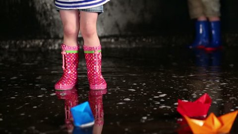 Girl in pink rubber boots jumping on puddle near colored paper boats