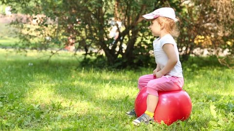 Smiling little girl on red ball for jumping at grass in park
