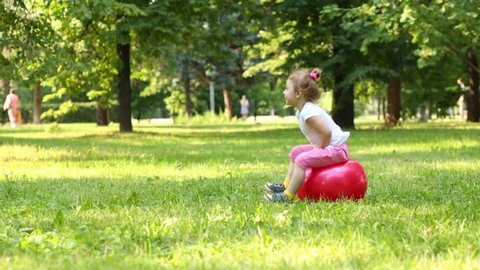 Cute little girl jumping on red ball at green lawn in park