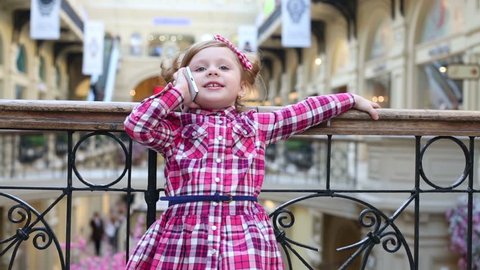 Little cute girl near wrought railings talks to mobile phone and runs at mall