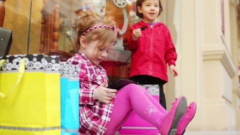 Little girl sits on floor with tablet pc and boy whistles near showcase with antiques at mall