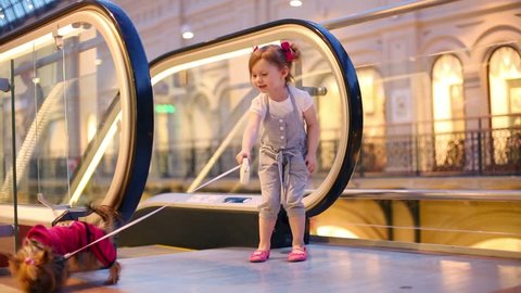 Little girl with small dog on leash stands next to escalator in mall