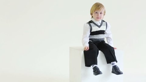 Little blond boy in vest sits on big cube, smiles and dangle feet