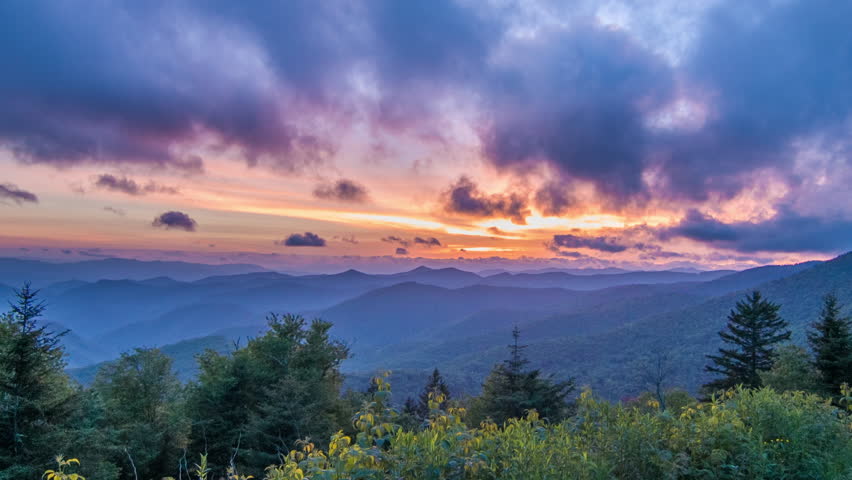 Vibrant Blue Ridge Mountain Sunset seen from Caney Fork Overlook on the Blue Ridge Parkway Featuring Green Forestry, Blue Layers of Mountains and Glowing Orange and Pink Sky near Asheville, NC.