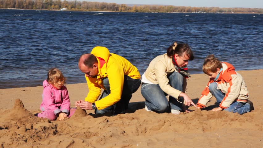 family of mother, father, son and daughter build hills of sand on river beach 