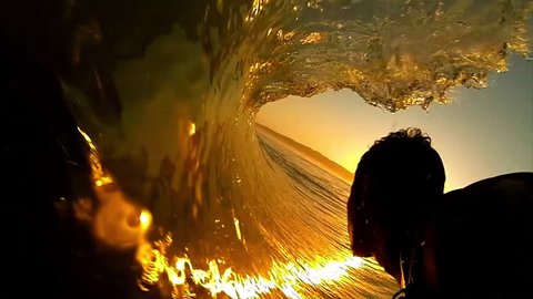High contrast POV shot of silhouetted surfer as he rides through the barrel of a dark orange wave, with the sun setting ahead of him