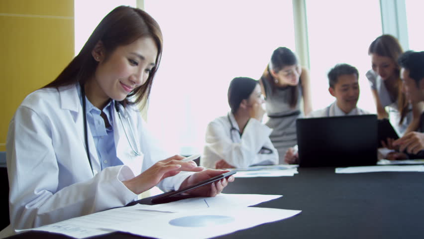 Young ethnic medical executives meeting in the boardroom of a modern hospital building female doctor white coat using mini tablet foreground
