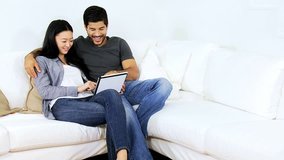 Smiling young Asian Chinese heterosexual couple sitting together home sofa following social media using touch screen tablet slow motion