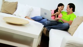 Young heterosexual Asian Chinese couple relaxing white home couch talking friends wireless laptop computer video technology