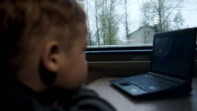 Close-up side view of a boy watching movie or cartoon on laptop during his traveling in the train. Focus on the window with passing trees and houses
