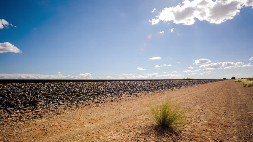 Timelapse, right to left at 45 degree angle up of a typical Karoo landscape with railway lines in a barren landscape with sun flare against a blue sky with scattered cumulus clouds. South Africa.