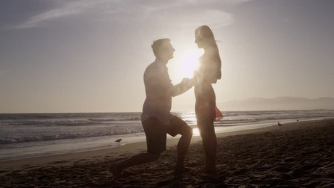 Romantic Silhouette of Man Getting Down on his Knee and Proposing to Woman on Beach - Couple Gets Engaged at Sunset - Man Putting Ring on Girl's Finger