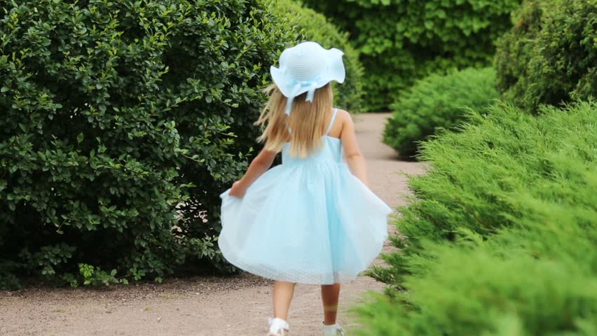 Happy little cute girl in blue dress and hat walks and turns near bushes in summer park