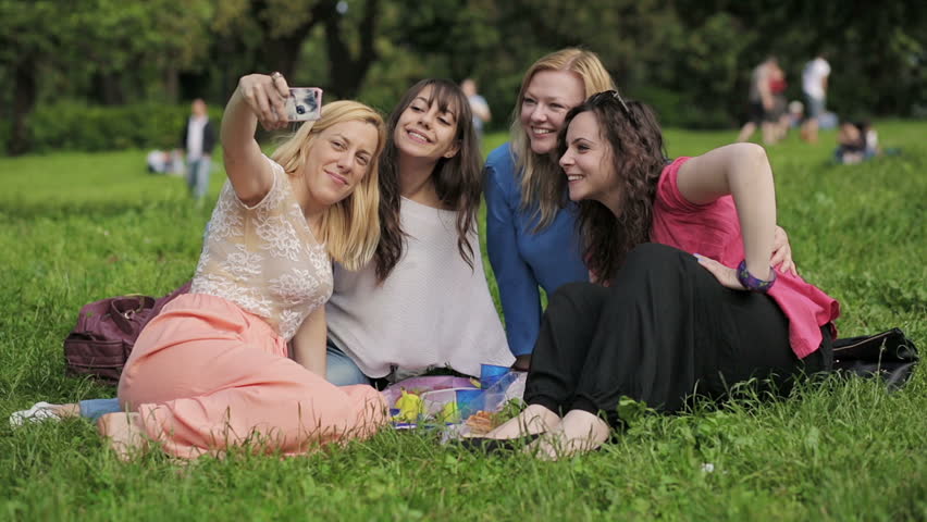 four cute girls taking photos of themselves oudoors on grass at picnic and laughing together: joy, happiness, outdoor, park, green. Selfies with friends!