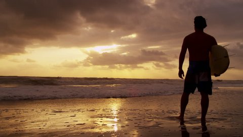 Silhouette solitary young male surfer standing sand beach sunset holding surfboard watching ocean waves shot on RED EPIC Vídeo Stock
