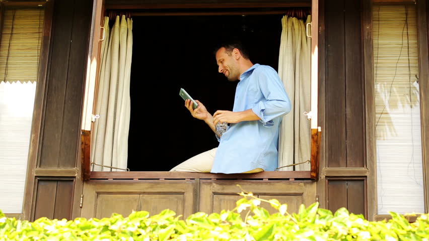 Young man with smartphone, drinking water in country house window
