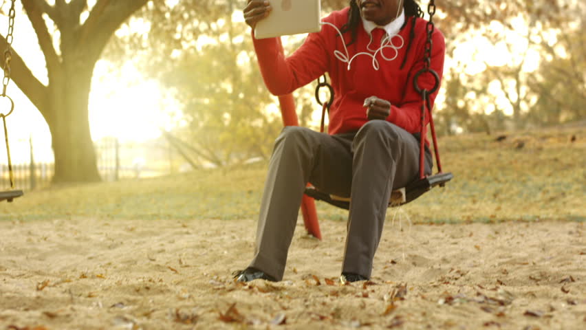 Panning shot of African American Man using tablet computer for  a camera while on the swing at local park at sunrise. 